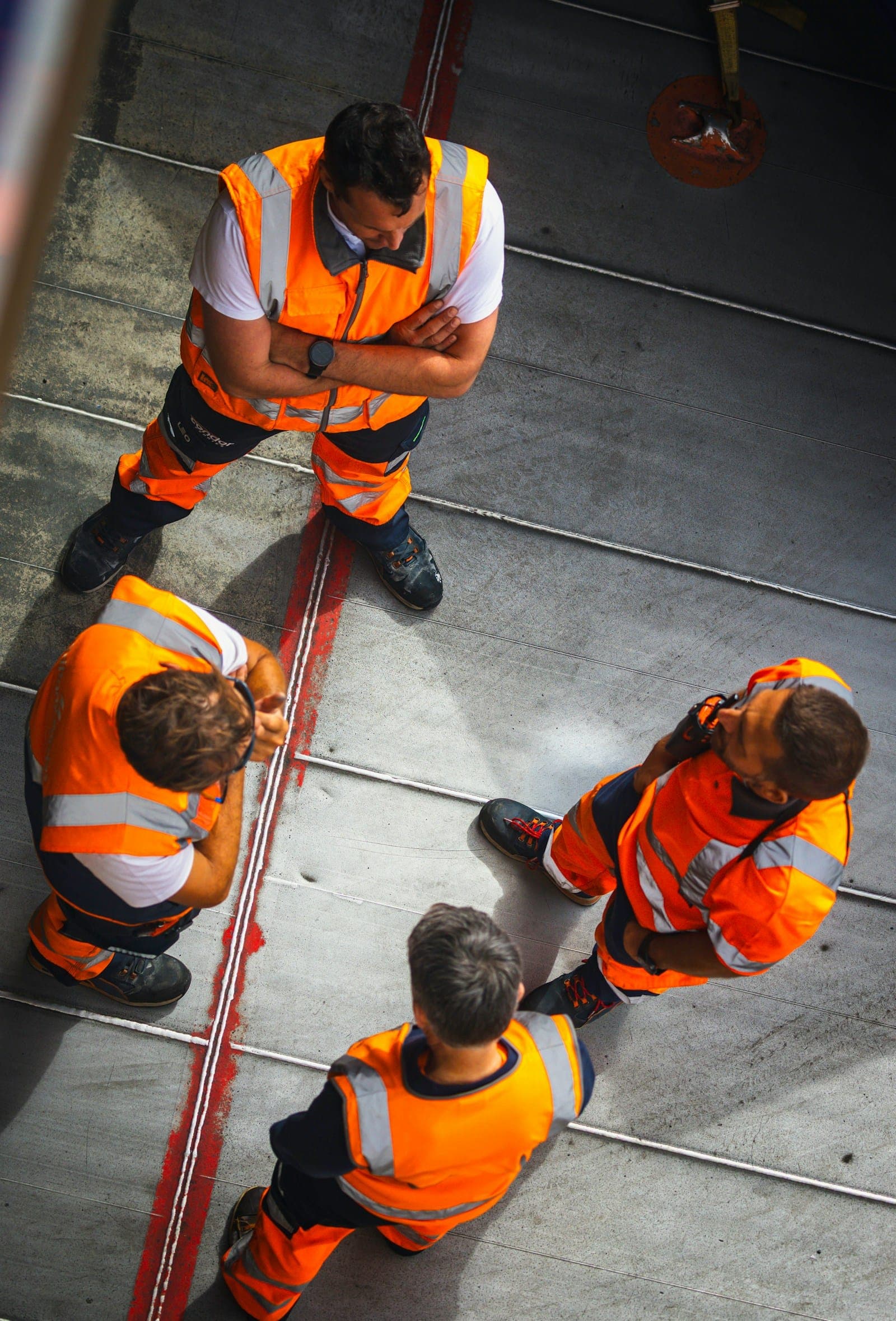 Four workers in orange hi-vis gather in a loose huddle before the start of a shift, seen from above.