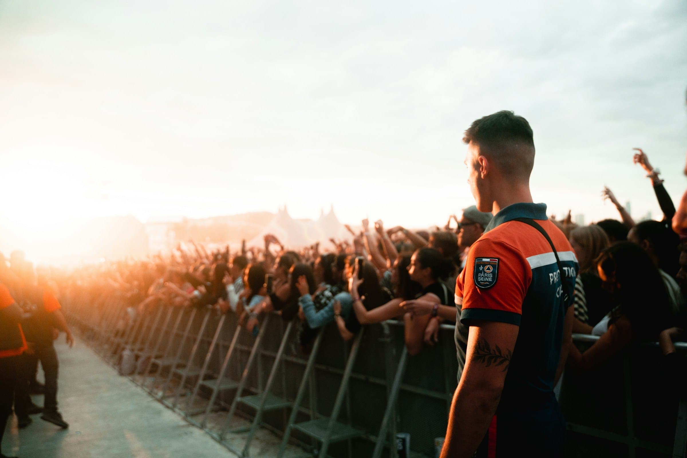 A steward stands at the front barrier of a packed festival crowd, watching fans with raised arms in golden evening light.