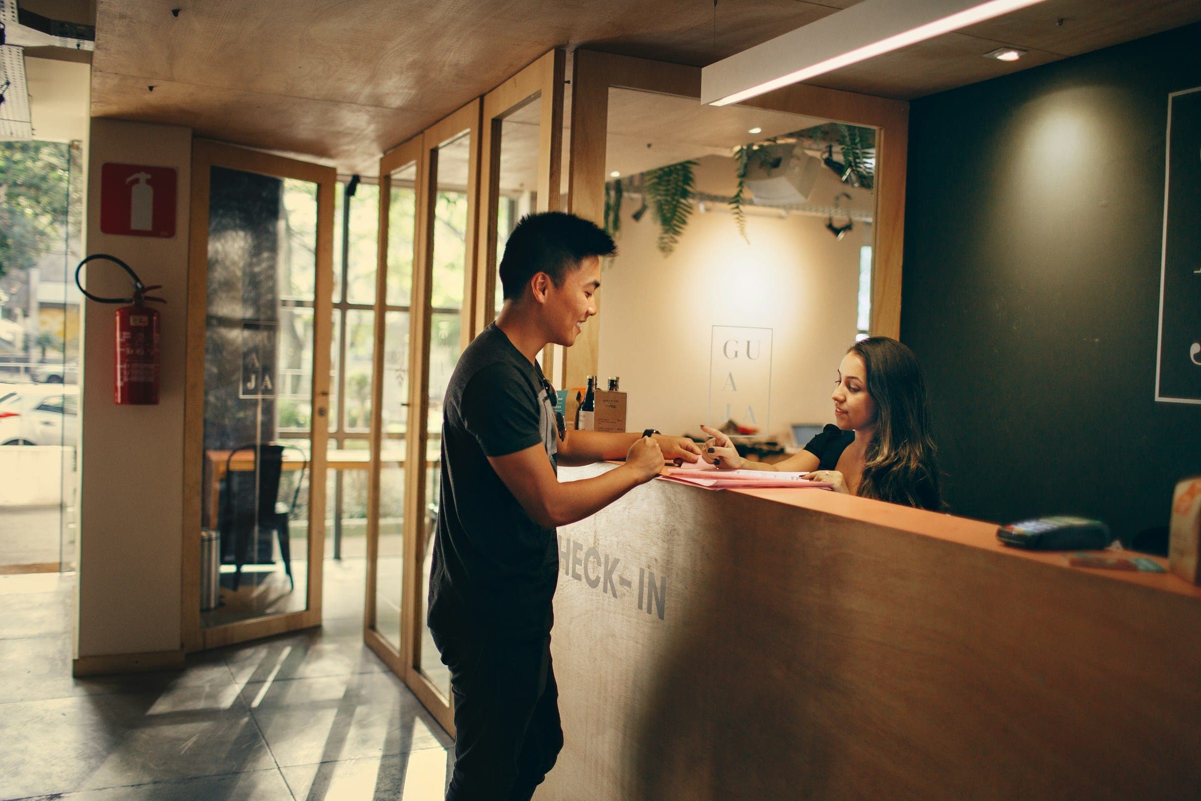 A visitor leans on a wooden check-in counter while a receptionist behind it helps him with paperwork.