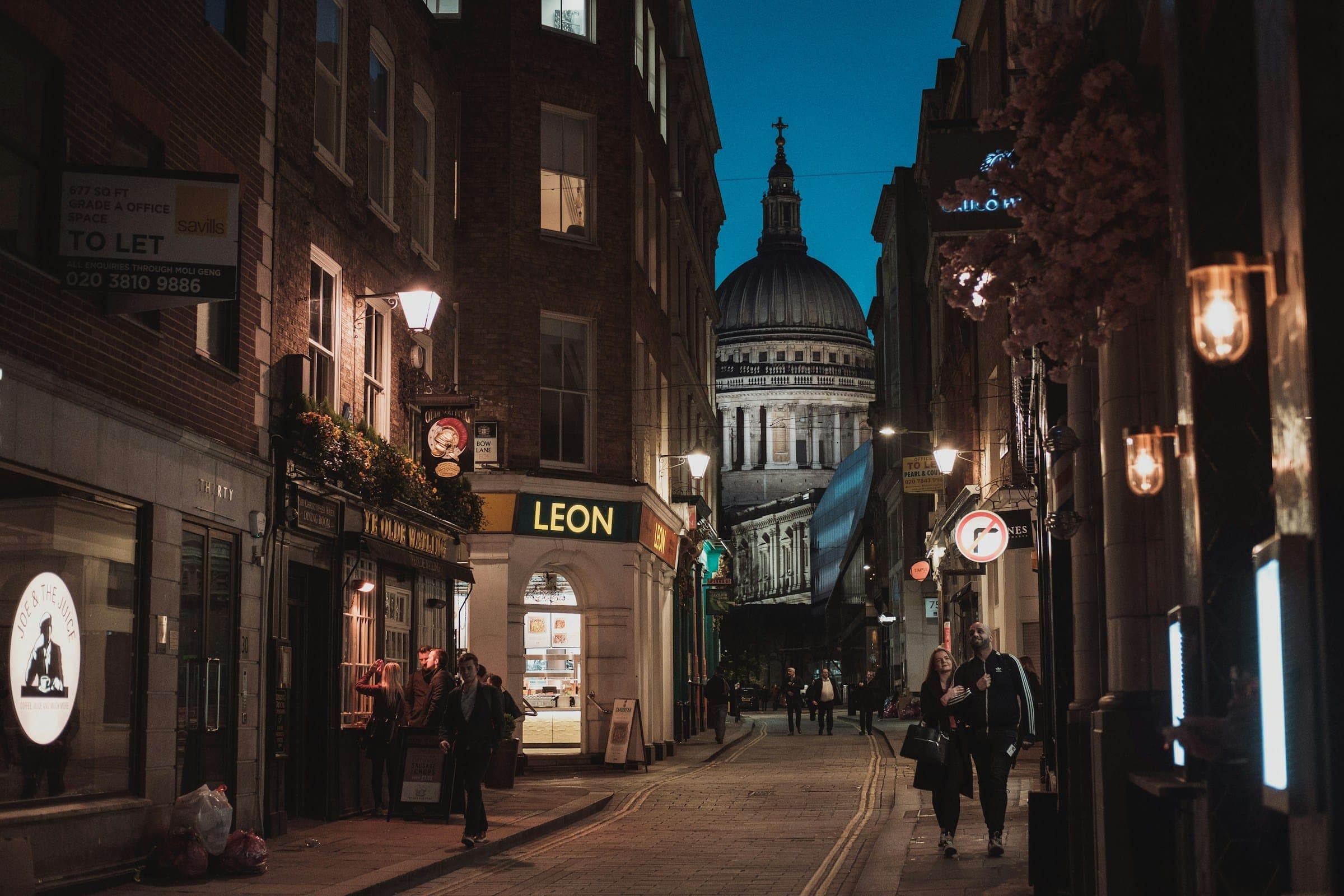 A narrow London street of shopfronts at dusk, warm wall lamps lit and the dome of St Paul's Cathedral against a deep blue sky.